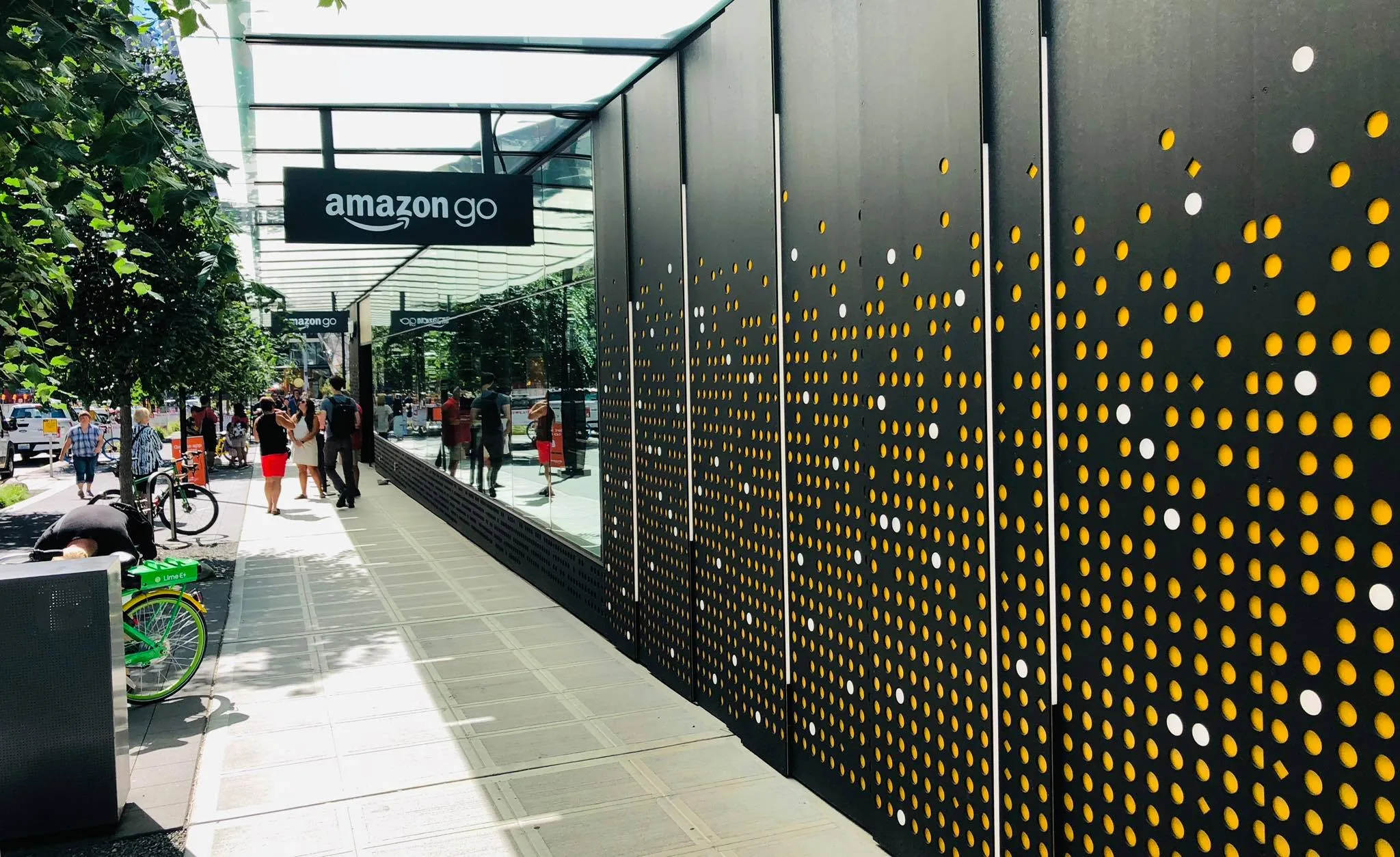 The Amazon Go storefront along a tree-lined sidewalk, its sign hanging under the awning as people walk past.