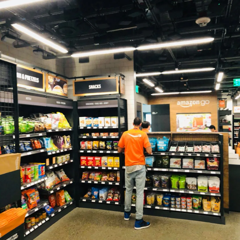 An Amazon Go employee restocking the snacks aisle, shelves stacked with chips, crackers and trail mix.