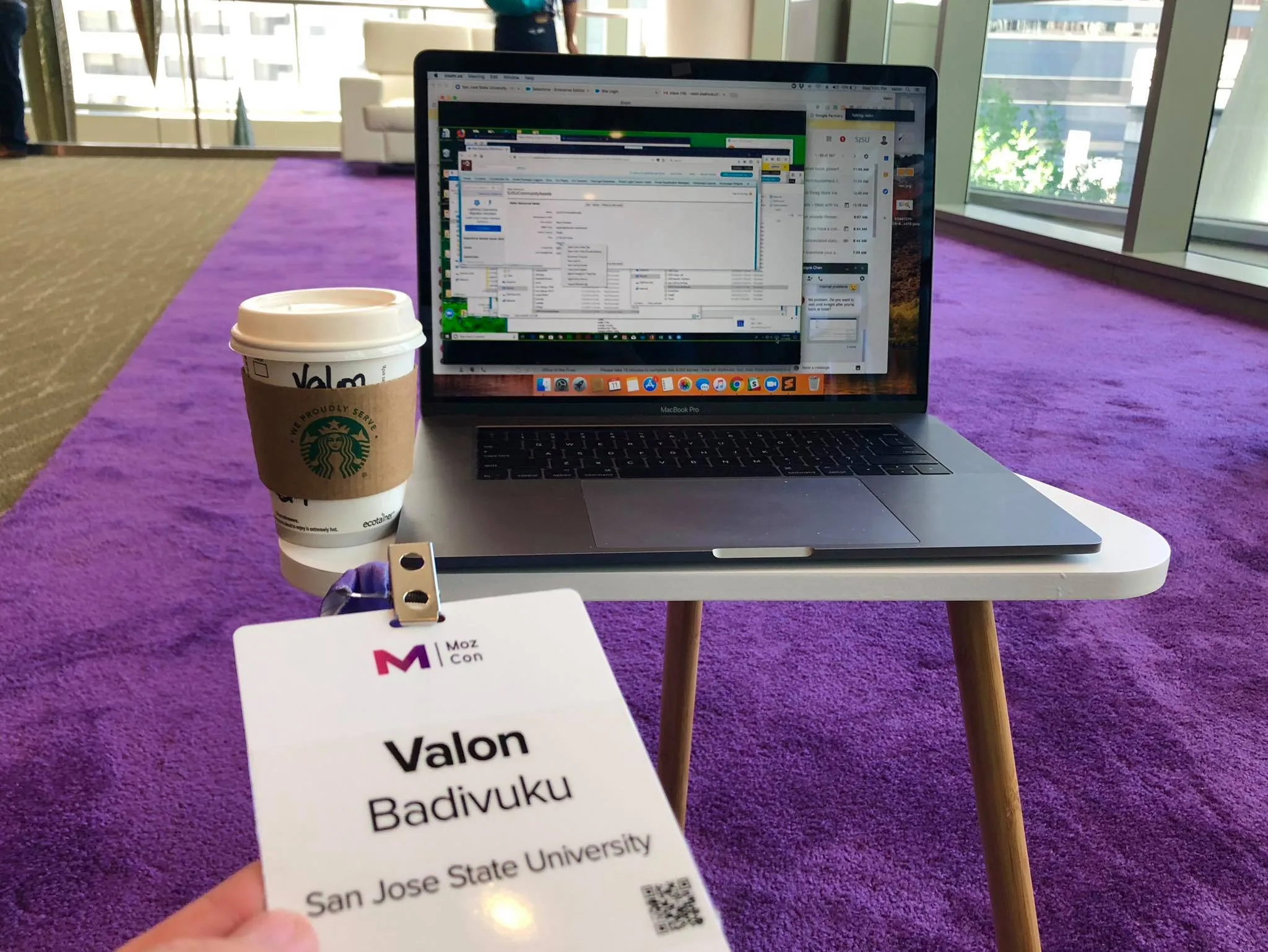 My MozCon badge, listing San Jose State University, propped against a MacBook Pro next to a Starbucks coffee on a small table with purple conference carpet in the background.