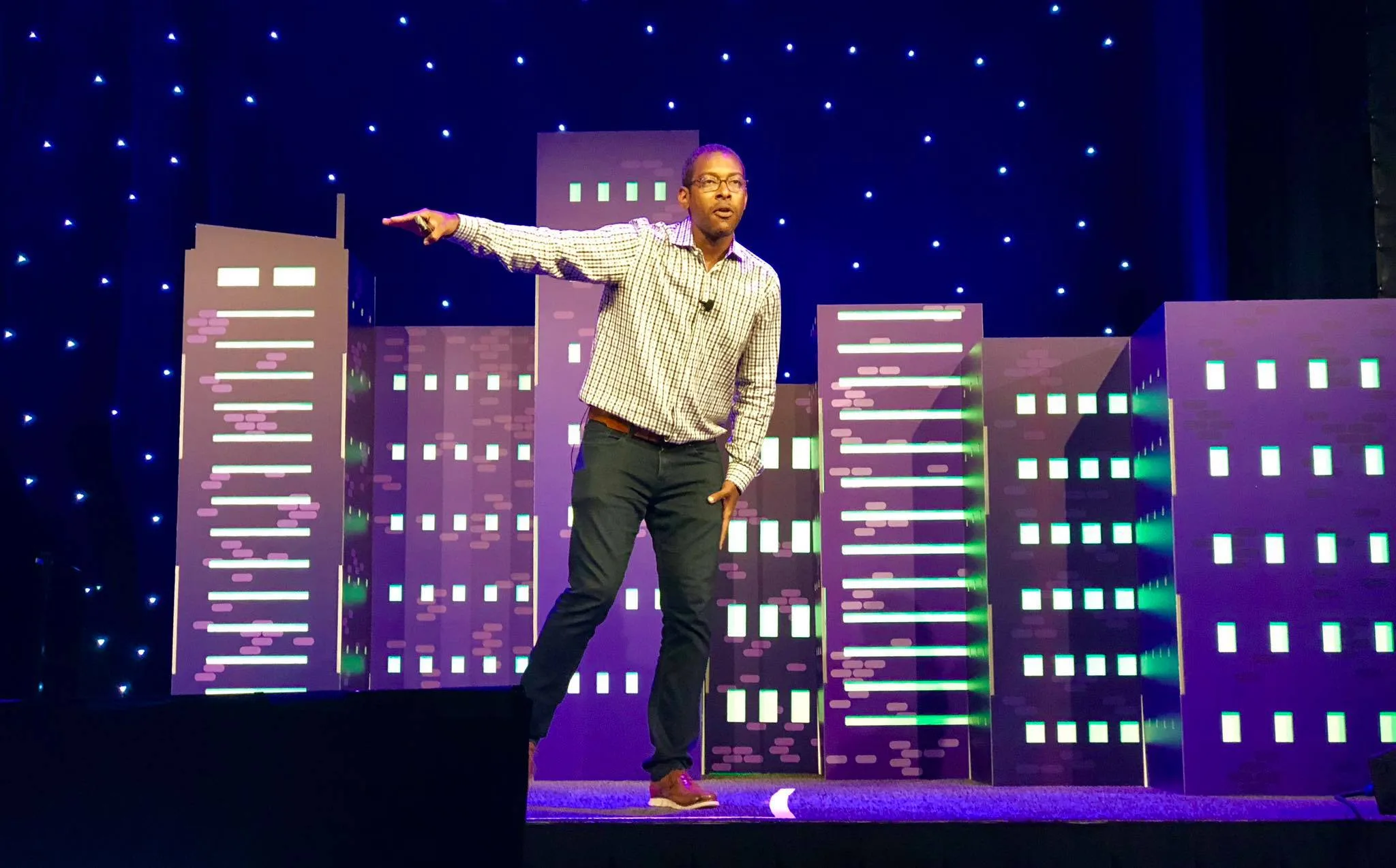 Michael King on the MozCon stage, gesturing mid-talk in front of a purple skyline backdrop under a starlit ceiling.