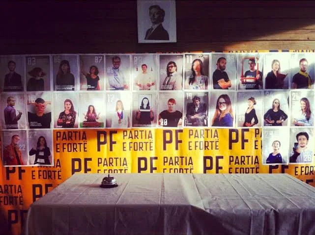 The wall of the Partia e Fortë campaign headquarters, papered with rows of numbered candidate posters above a run of orange PF banners, an empty table with a service bell in front of it.