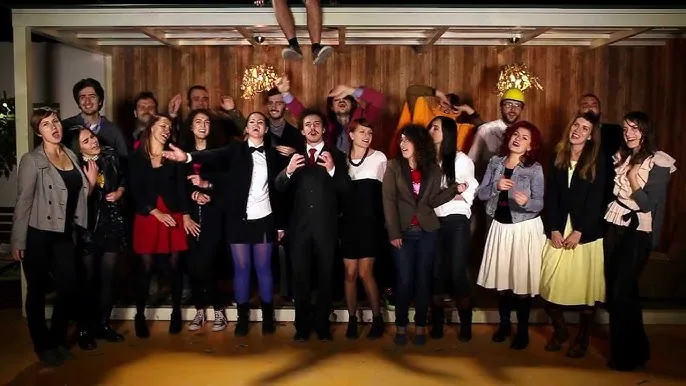 A still from the "Do të bëhem milioner" music video: about twenty people crowded together singing to camera, a man in a dark suit and red tie front and centre and a pair of legs dangling from the ceiling above them.